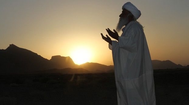 Betender Mann in weißem Gewand und Turban vor untergehender Sonne über Bergen