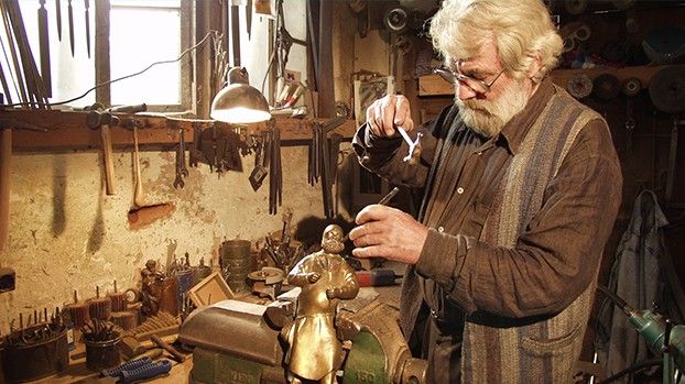 Älterer Mann mit Bart bearbeitet eine Bronzefigur in einer Werkstatt voller Werkzeuge